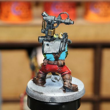 A painted figurine of a character with a large headpiece and shorts, holding a tool.